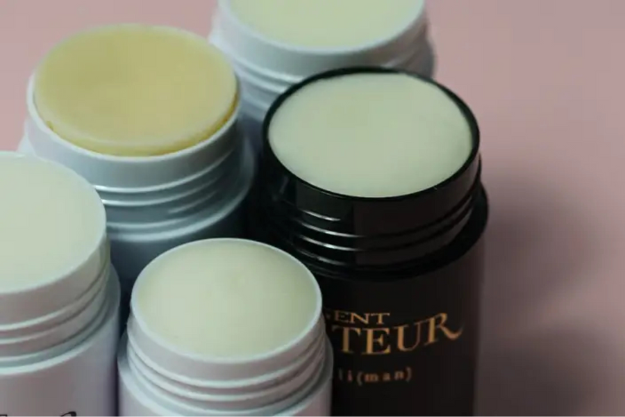 Open jar of balm, a pot of cream deodorant and an uncapped stick with eucalyptus leaves on a pale blush surface